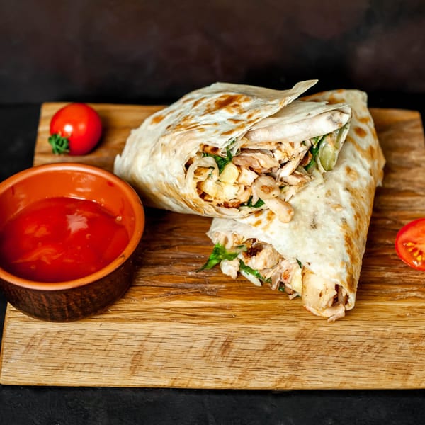 Two chicken shawarma wraps with a side of red sauce and cherry tomatoes on a wooden board.