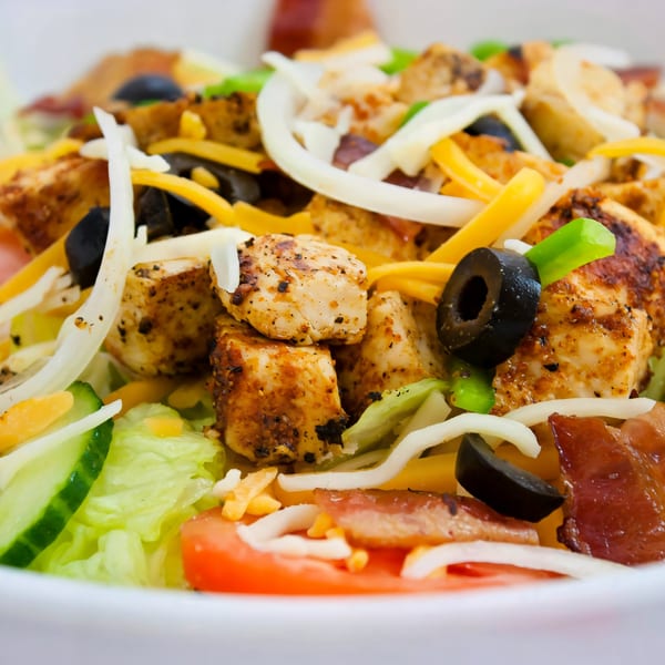 Close-up of a vibrant salad with grilled chicken, black olives, cheese, and fresh vegetables.