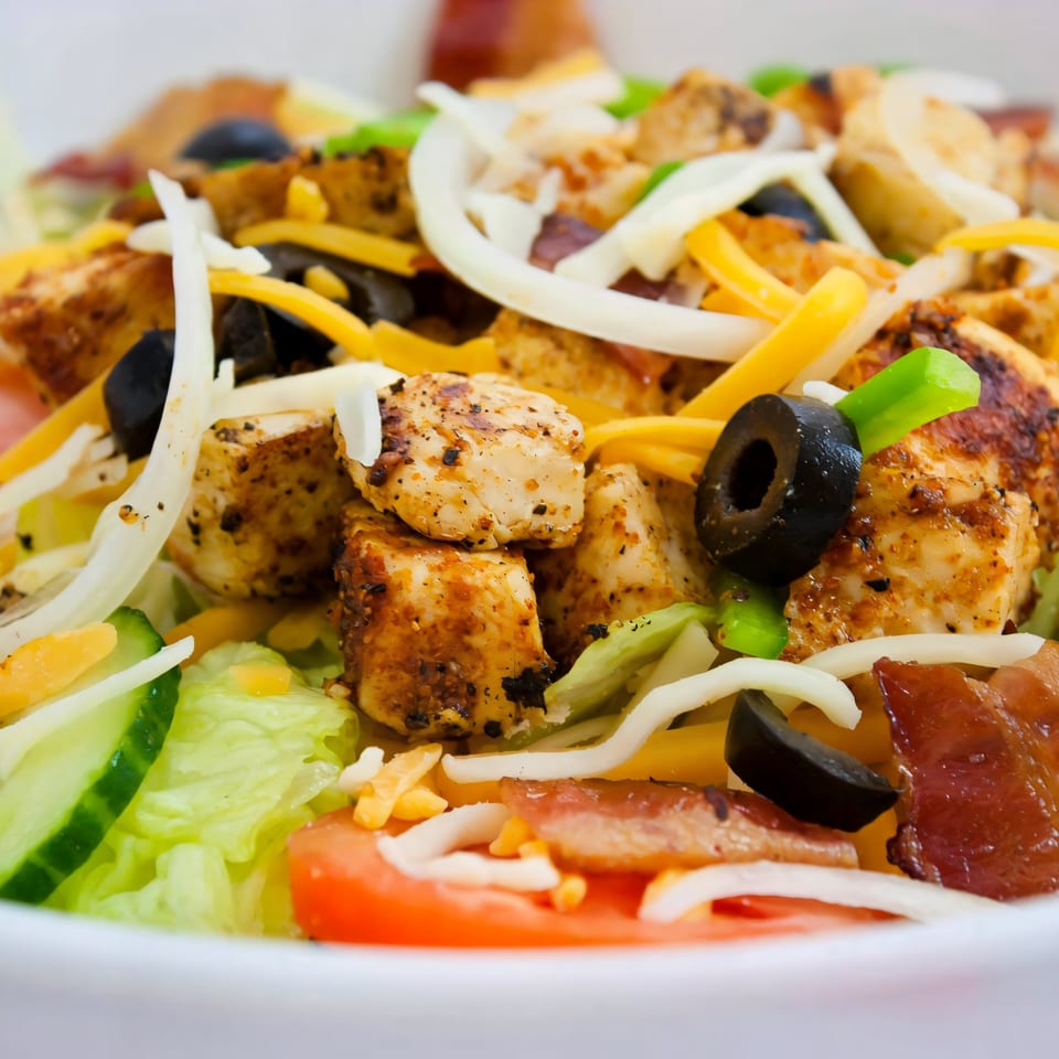 Close-up of a vibrant salad with grilled chicken, black olives, cheese, and fresh vegetables.